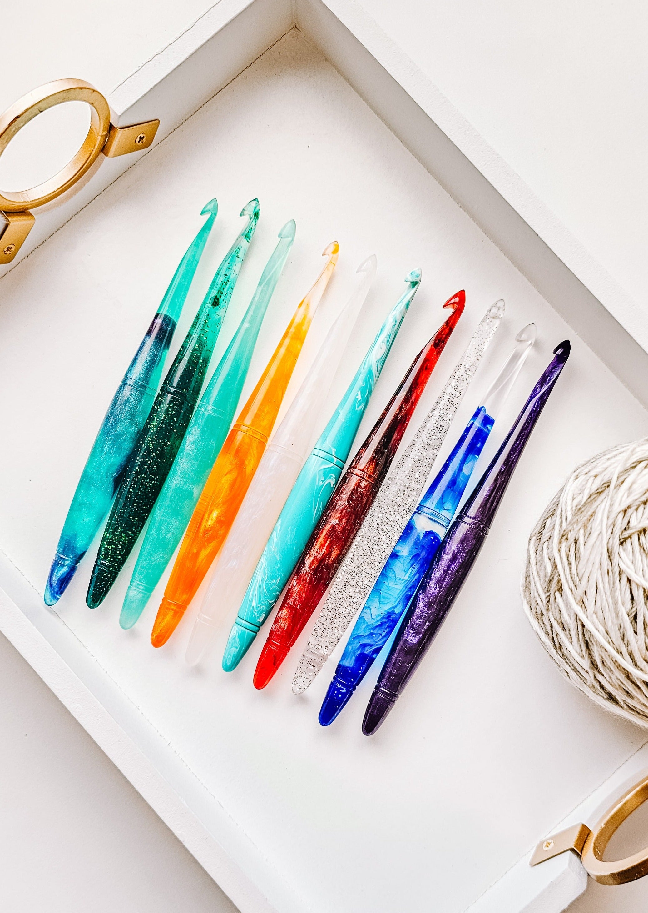 10 crochet hooks sit on a tray next to a ball of yarn. The tray is white.